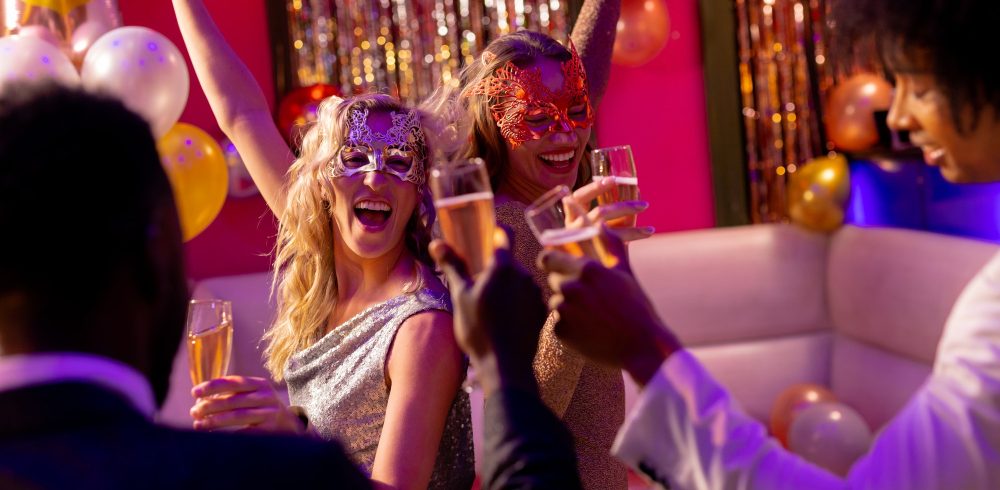 Two happy caucasian female friends in masks dancing and drinking champagne at a party in a nightclub