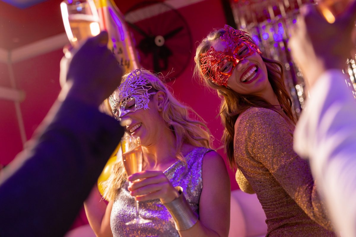 Two happy caucasian women wearing masks dancing and drinking champagne at a party in a nightclub